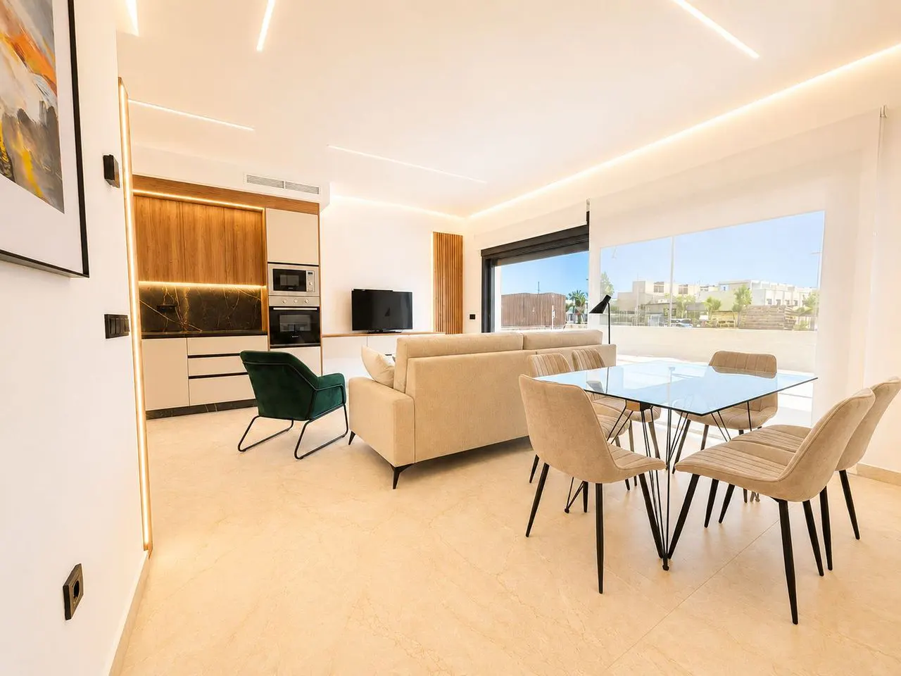 Bright, modern living space with a glass table, beige chairs, and a beige sofa. A green chair sits near a kitchen area with wood cabinets. Large window with a view.