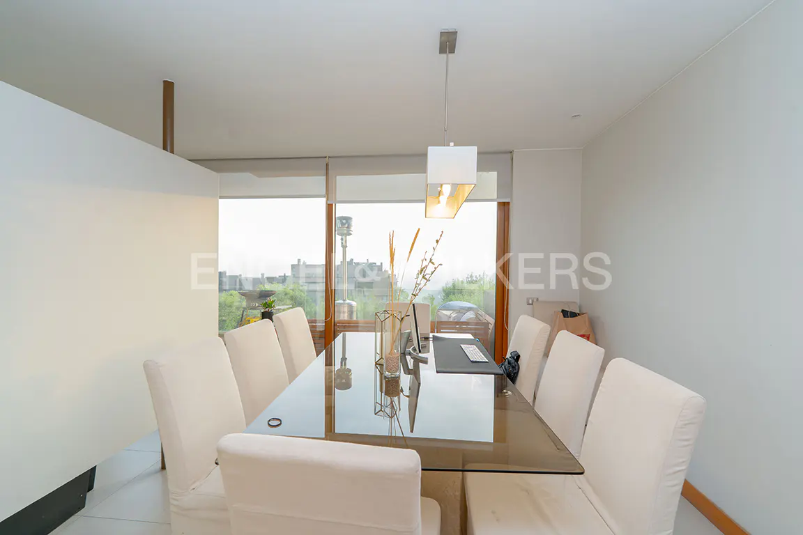 Bright dining room with a glass table, white chairs, and a modern light fixture. A large window offers a city view.