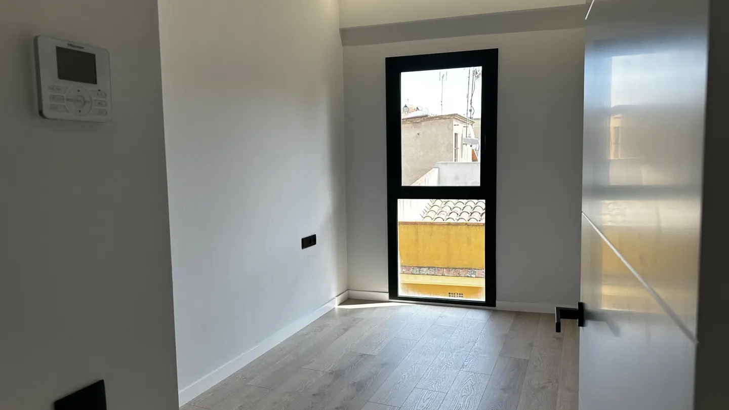 Bright, empty room with white walls, light wood floors, and a black-framed window showing a building exterior. A thermostat is on the left wall.