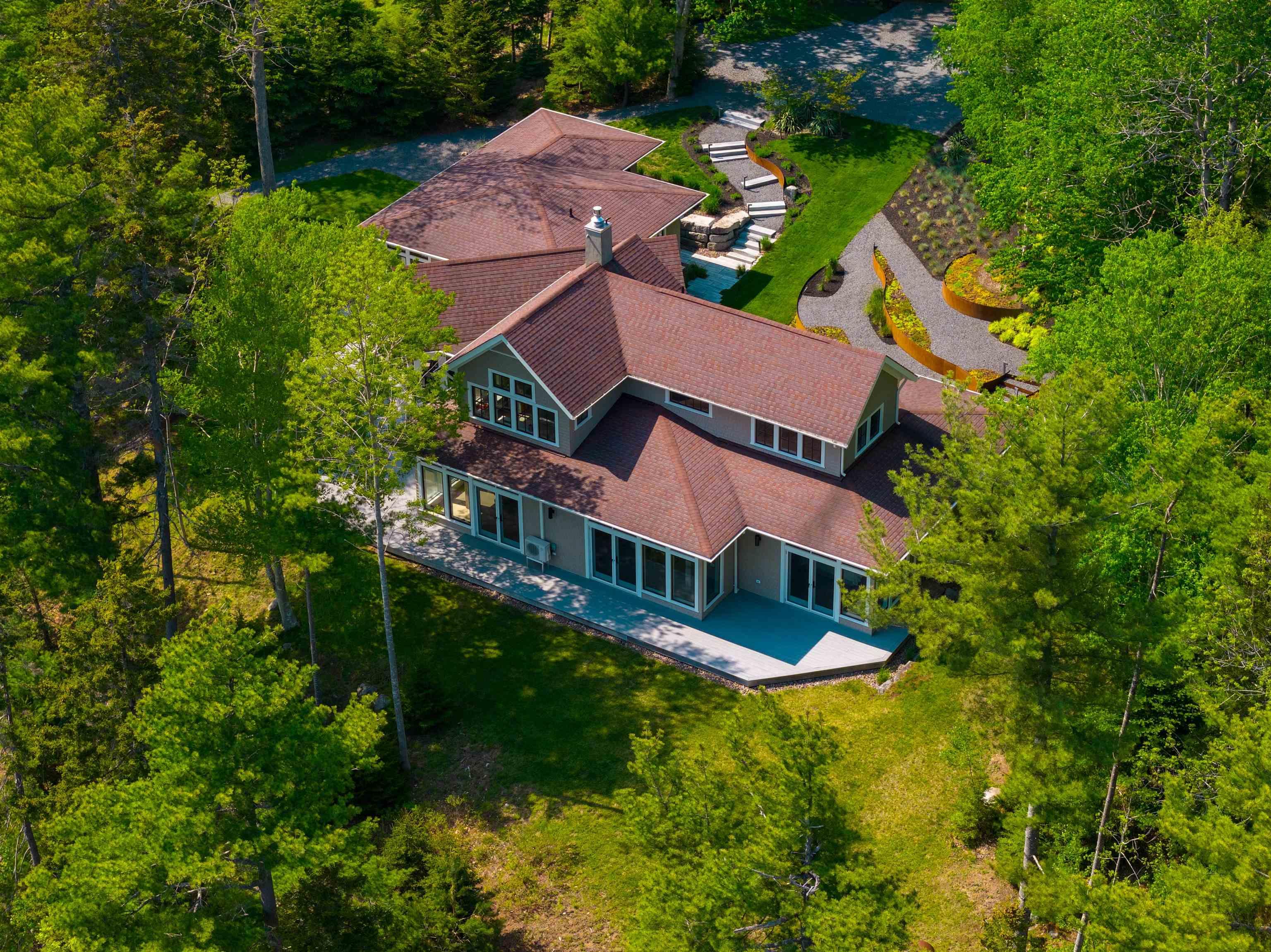 Aerial view of a tan house with a brown roof, surrounded by lush green trees, a stone walkway, and a landscaped yard.