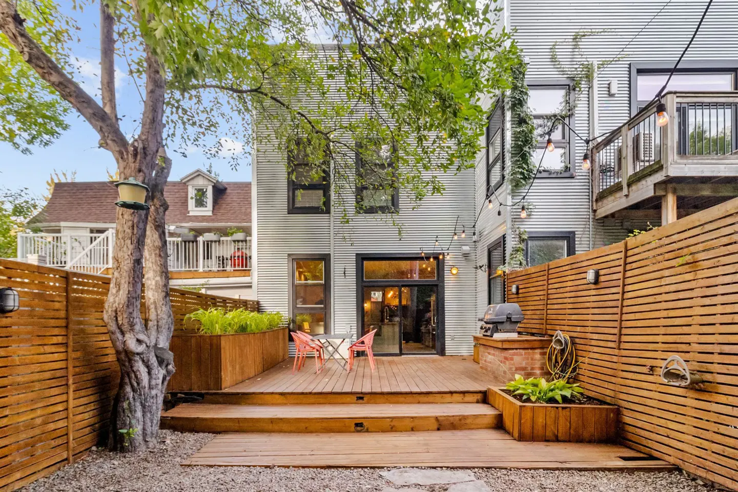 Backyard with a wooden deck, table, chairs, grill, and a corrugated metal house with string lights.