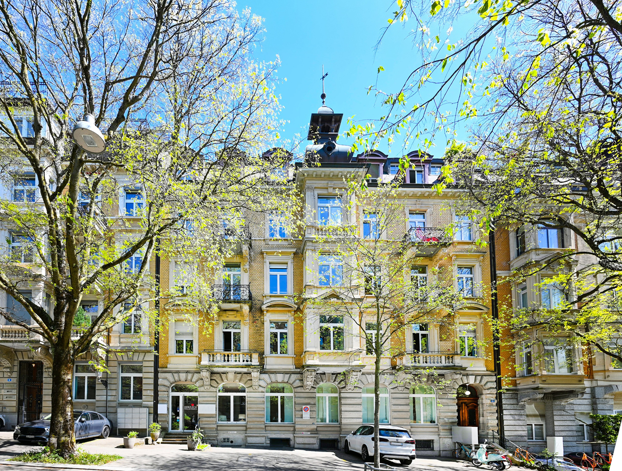 A yellow apartment building with blue windows is framed by trees with green leaves. Cars are parked on the street.