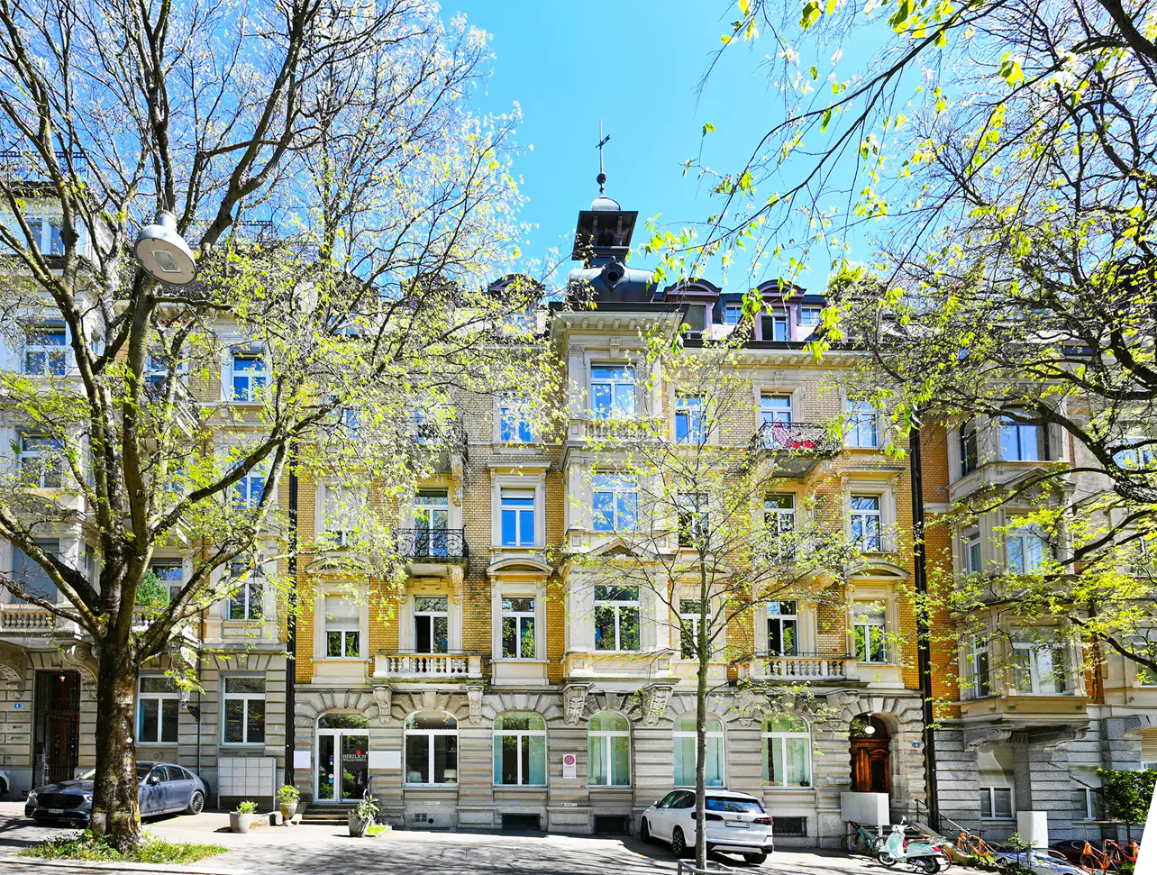 A yellow apartment building with blue windows is framed by trees with green leaves. Cars are parked on the street.