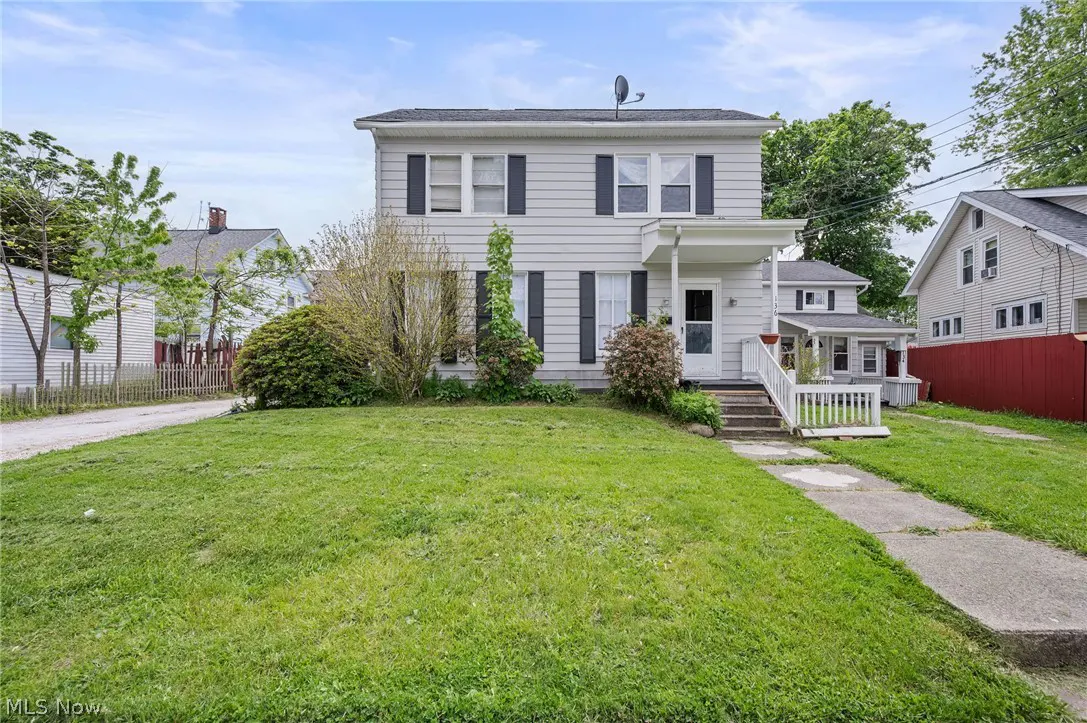 Two-story light gray house with black shutters, green lawn, and concrete walkway.