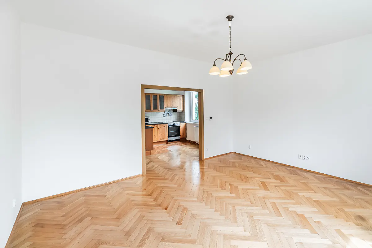Empty room with white walls, herringbone wood floor, and doorway to a kitchen with wood cabinets. A chandelier hangs from the ceiling.