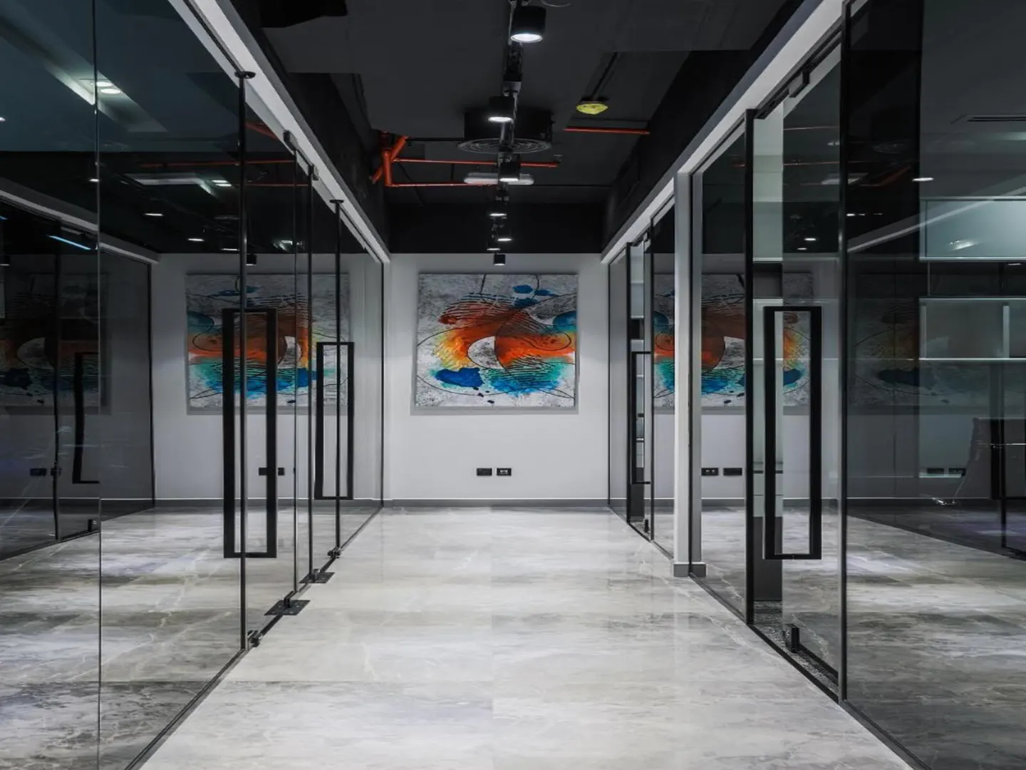 Office hallway with glass-walled offices, a gray marble floor, and abstract art on the back wall.