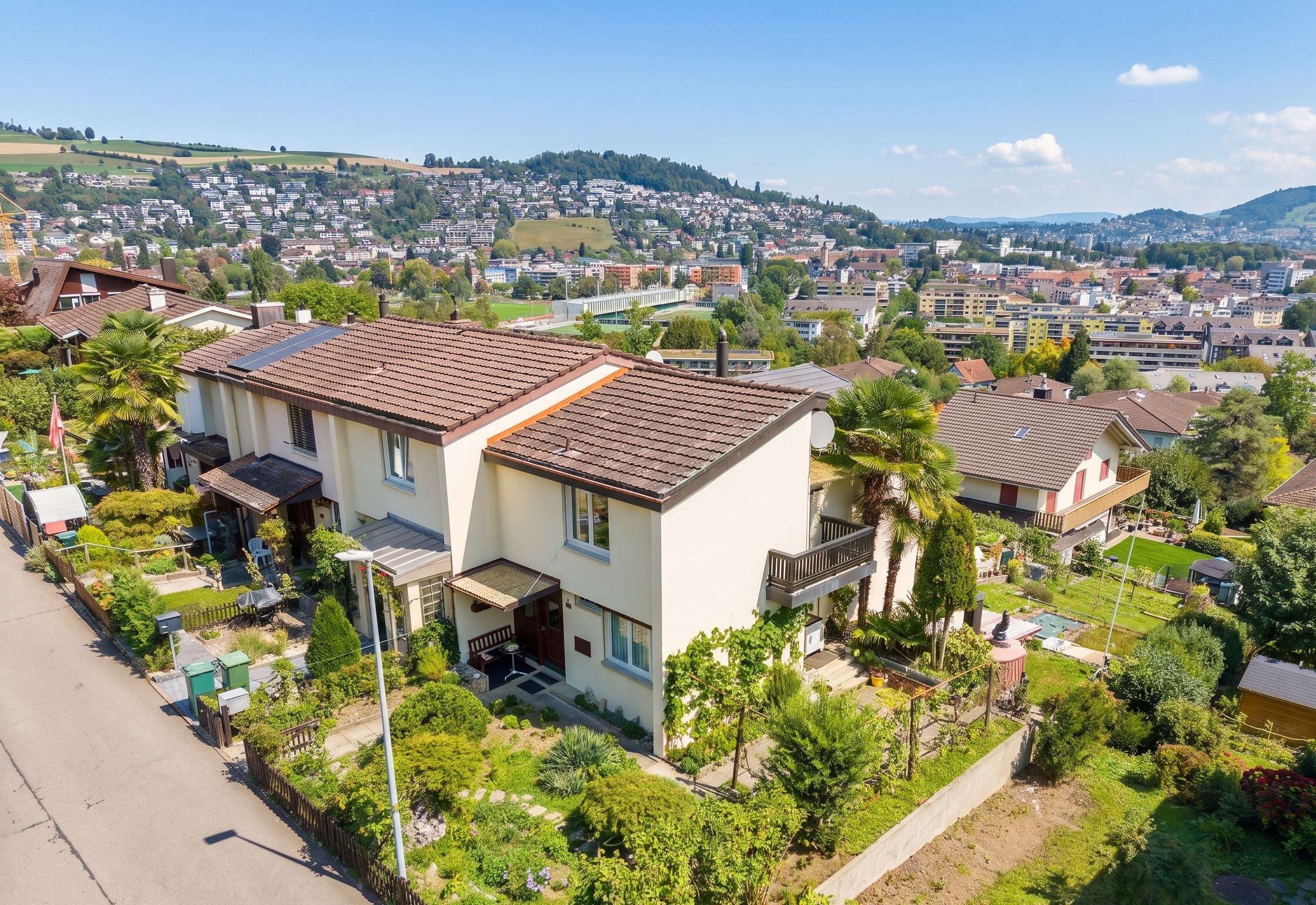 Aerial view of a cream-colored townhouse with a brown roof, lush garden, and palm trees, overlooking a hillside town. [AI-generated text]