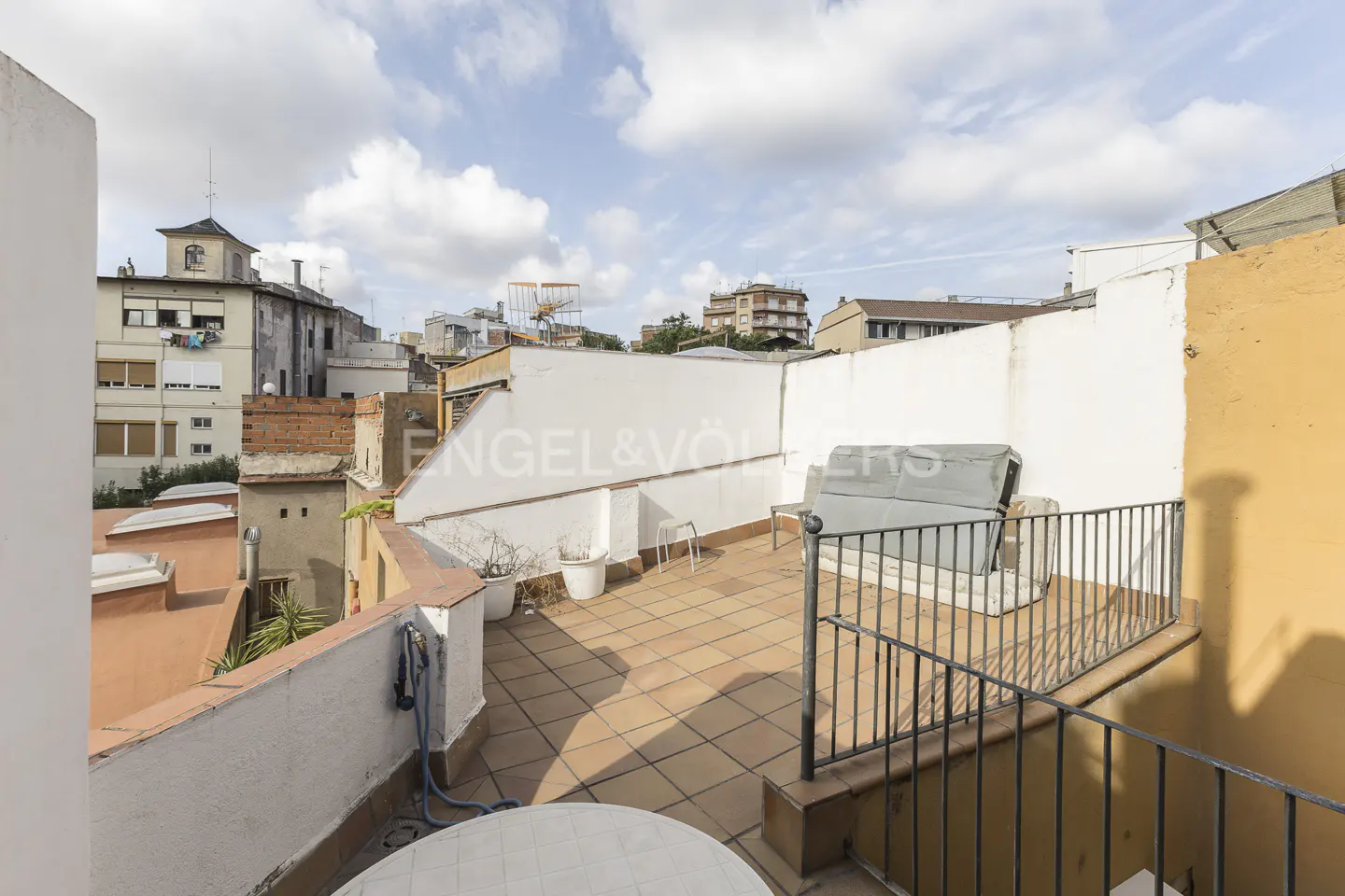 A rooftop terrace with brown tiles, white walls, and a black metal railing. A white table and chairs are visible. Buildings are in the background.