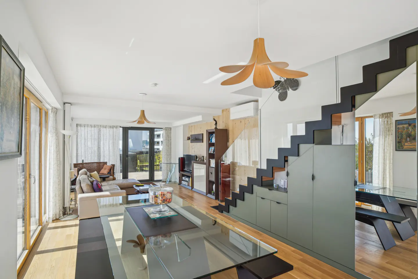 Bright, modern living room with glass table, wood floors, and black staircase. A beige sofa sits near large windows.