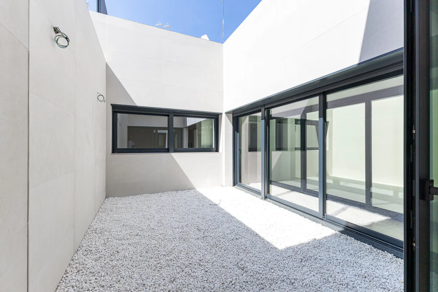 A modern courtyard with white walls, a black-framed window, and sliding glass doors. The ground is covered in white pebbles.