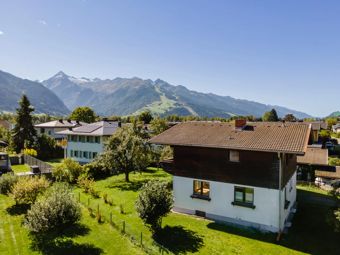 Two-story house with a brown roof and white walls, green lawn, and mountains in the background under a blue sky.
