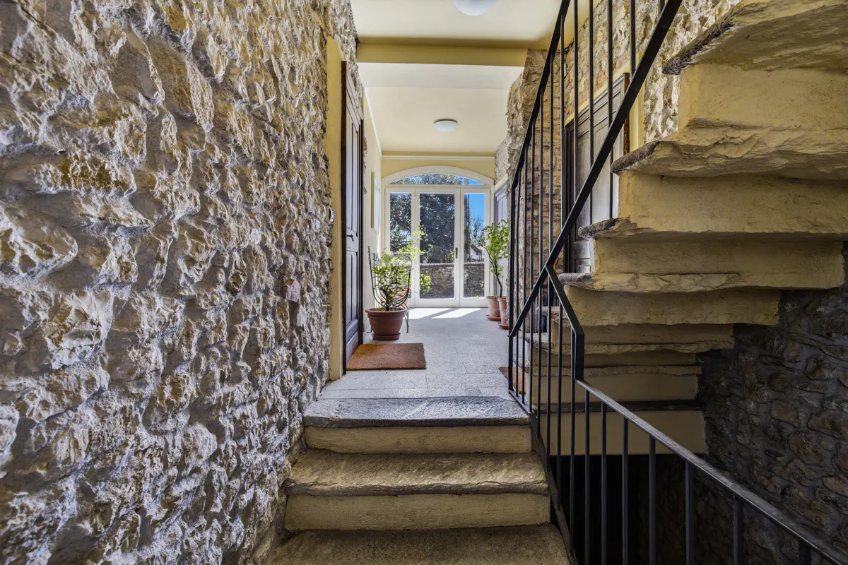 Stone hallway with stairs leading to a bright sunroom. Black metal railing on the right. Potted plants line the sunroom.