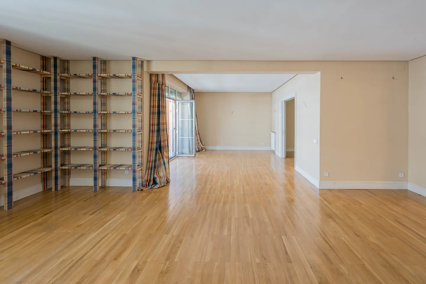 Empty room with hardwood floors, beige walls, and a built-in bookshelf with plaid fabric trim. Glass doors lead to a balcony.