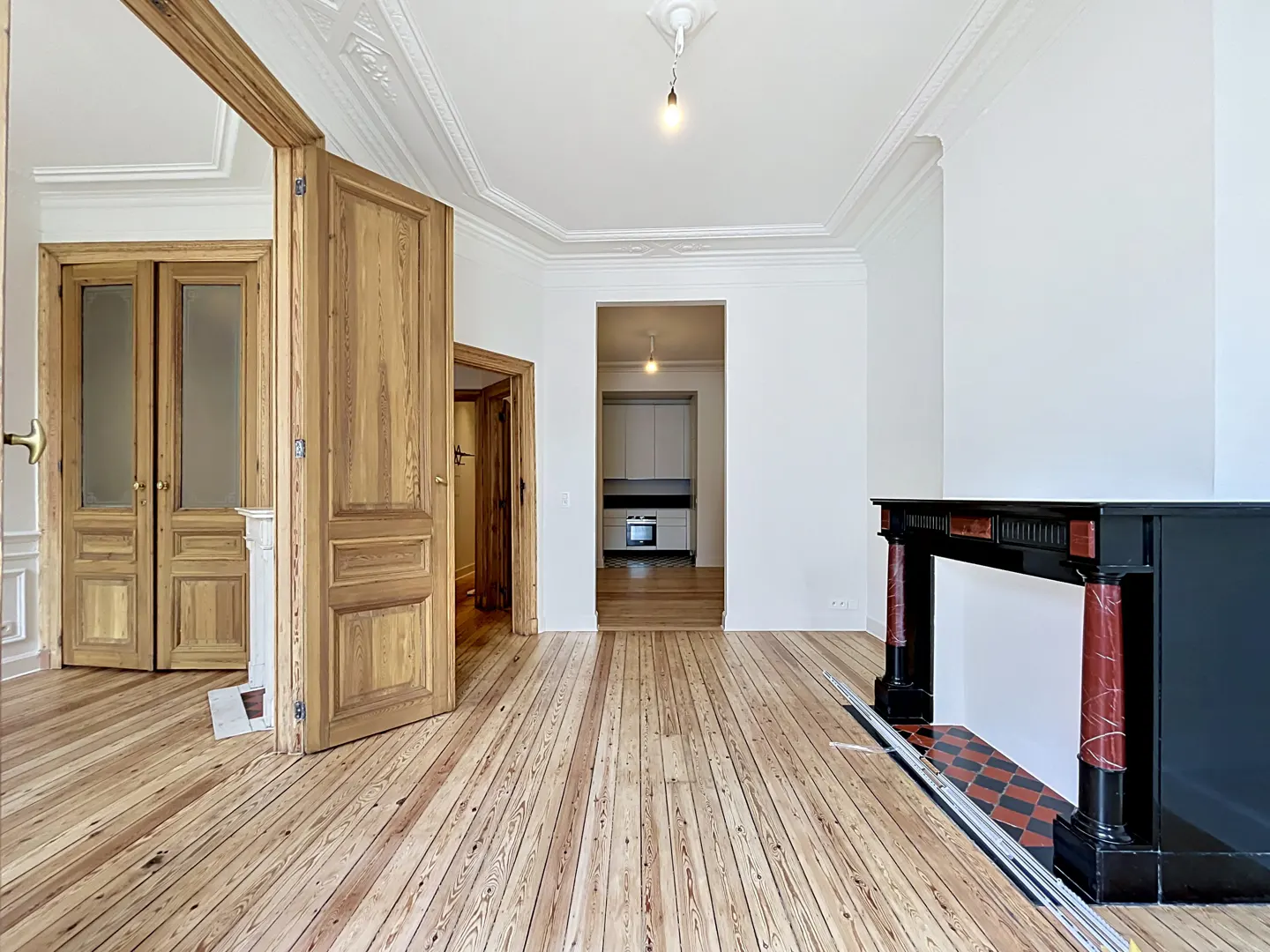 Bright room with wood floors, white walls, and ornate ceiling. A black fireplace is on the right, and doorways lead to other rooms.