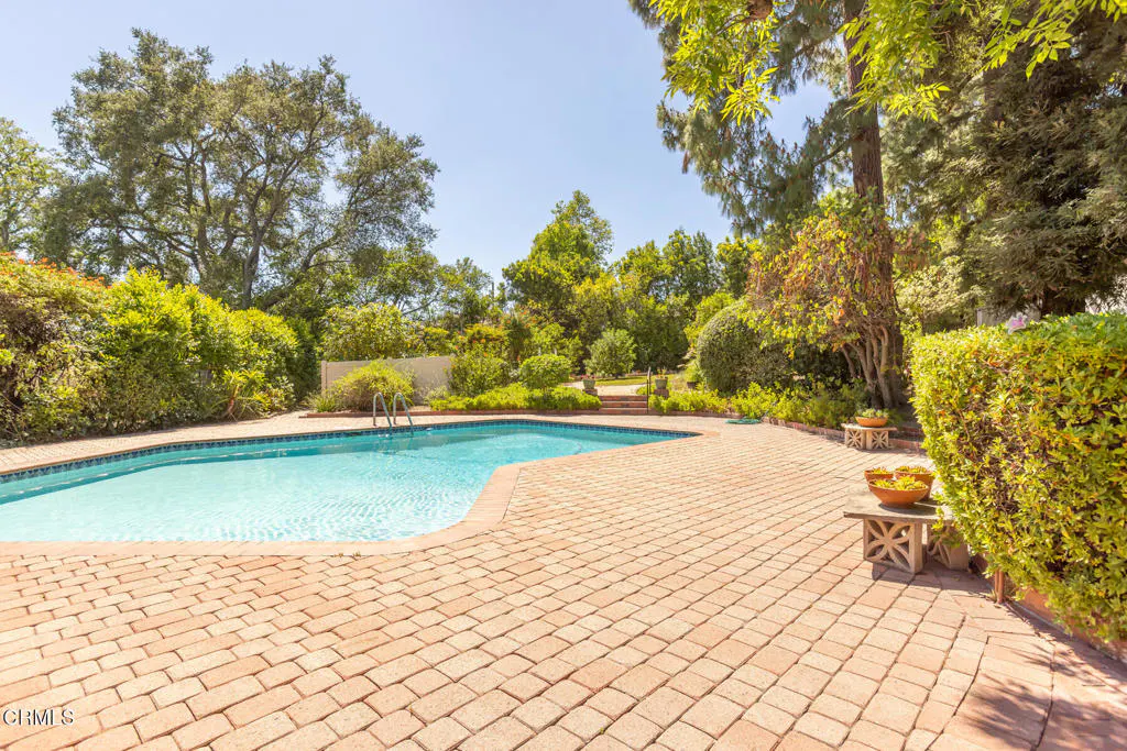 Outdoor pool with brick patio, surrounded by green trees and bushes under a blue sky.