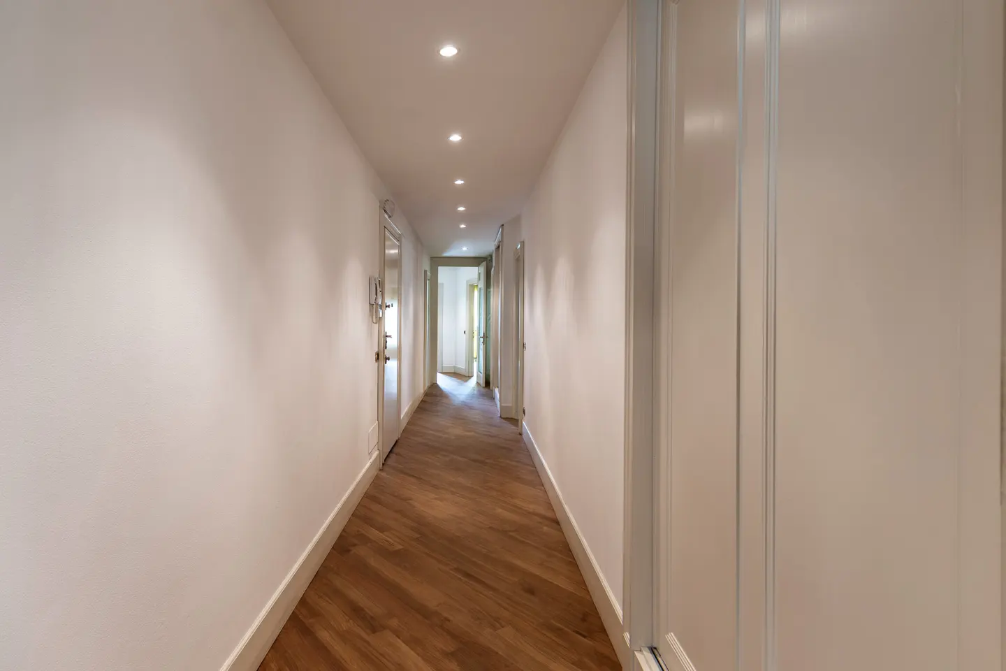 Bright hallway with white walls, wood floors in a herringbone pattern, and recessed lighting. Doors line the hall.