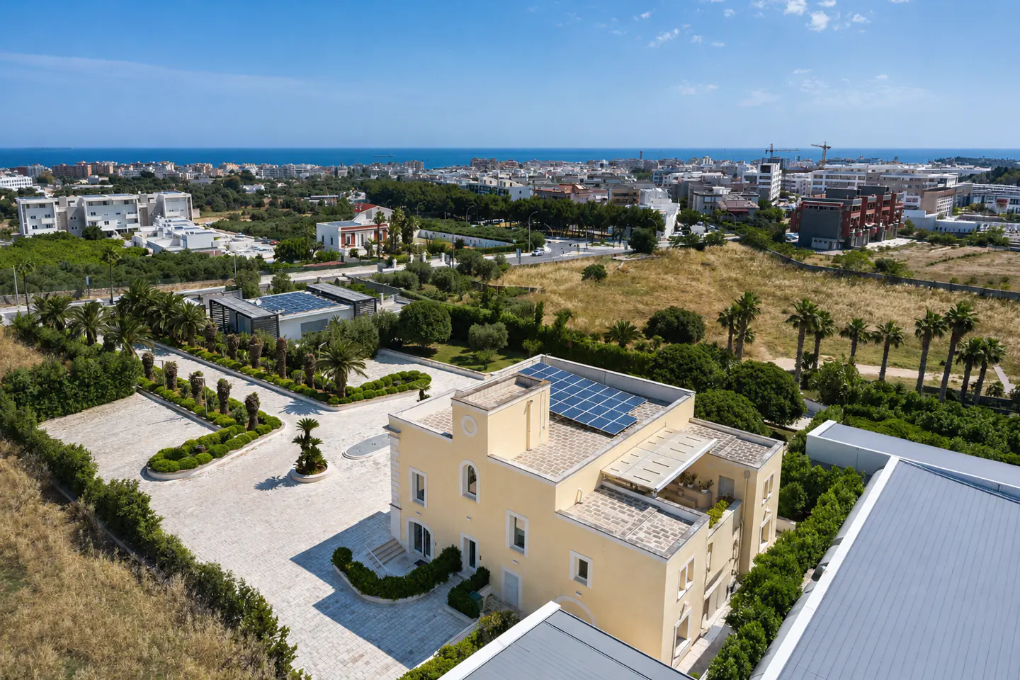 Aerial view of a yellow villa with solar panels, a paved courtyard, and lush gardens, overlooking a coastal city and sea.