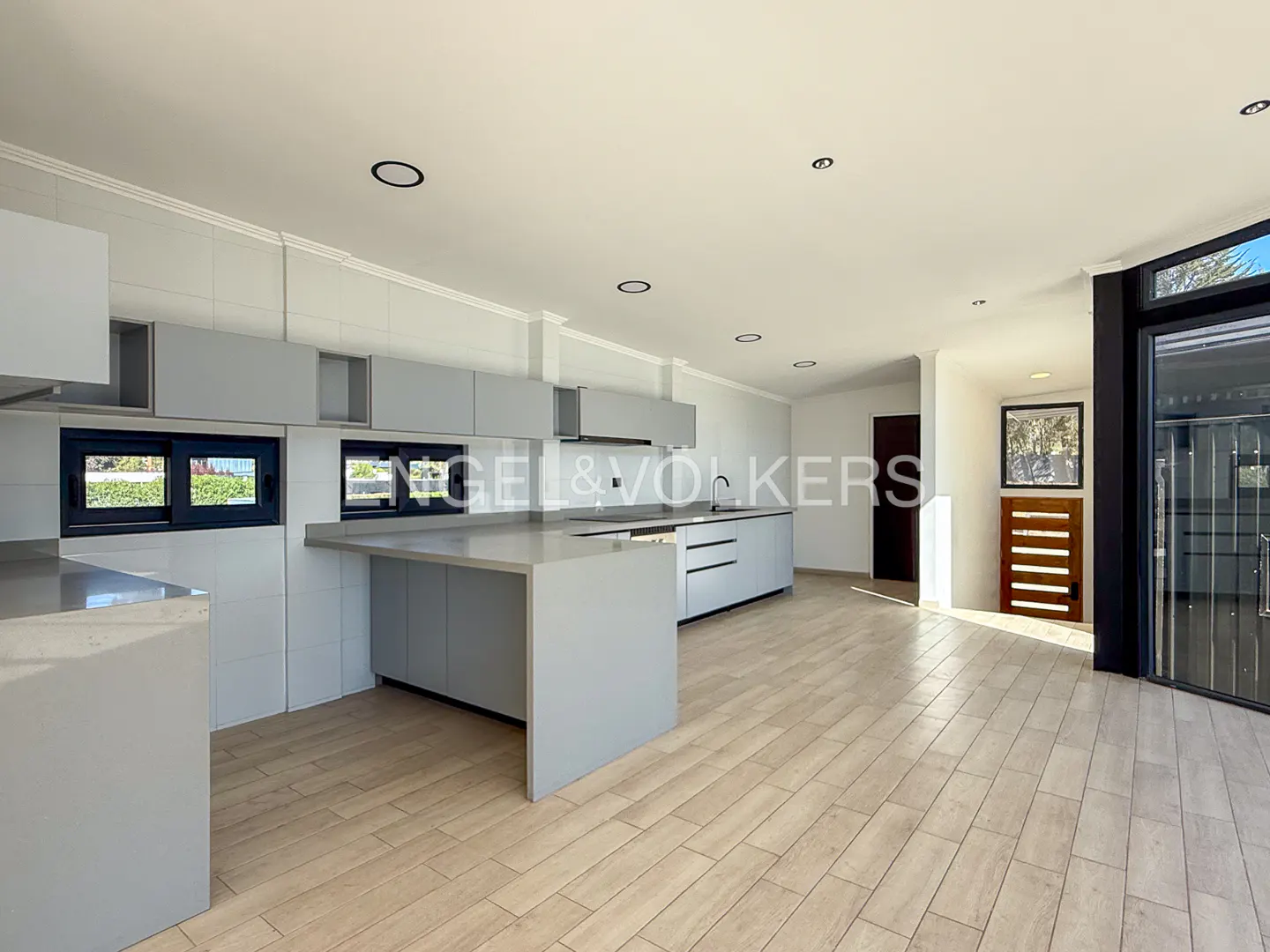 A bright, open kitchen with light gray cabinets, countertops, and wood-look tile flooring. Black framed windows and a sliding glass door provide natural light.