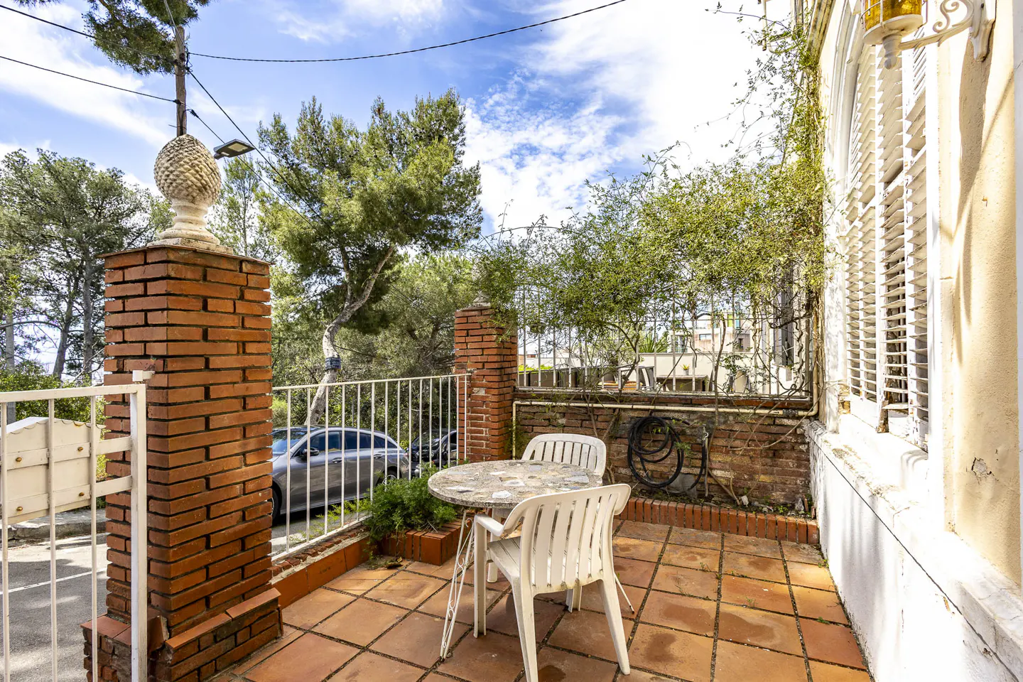 Outdoor patio with terracotta tiles, white plastic table and chairs, brick pillars, and a view of trees and a car.