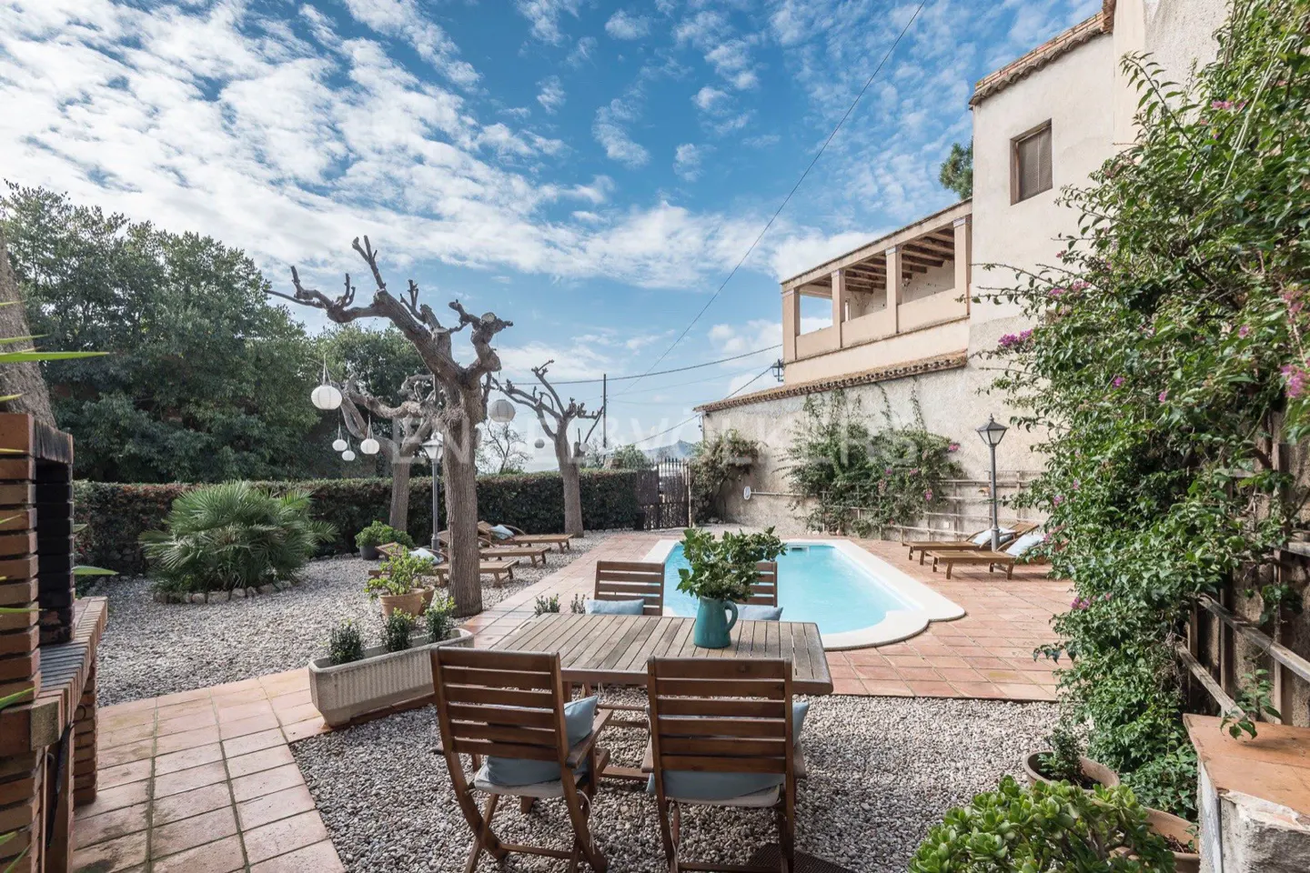 Outdoor patio with a pool, table, and chairs. The patio is surrounded by trees and plants, and the sky is blue with white clouds.