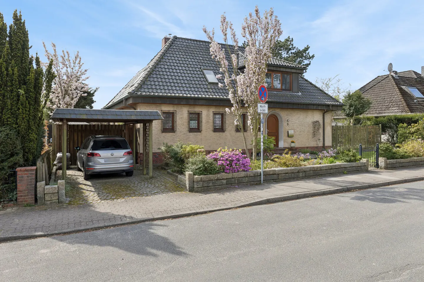 Two-story house with a gray car in the carport, a garden with purple flowers, and a gray roof.