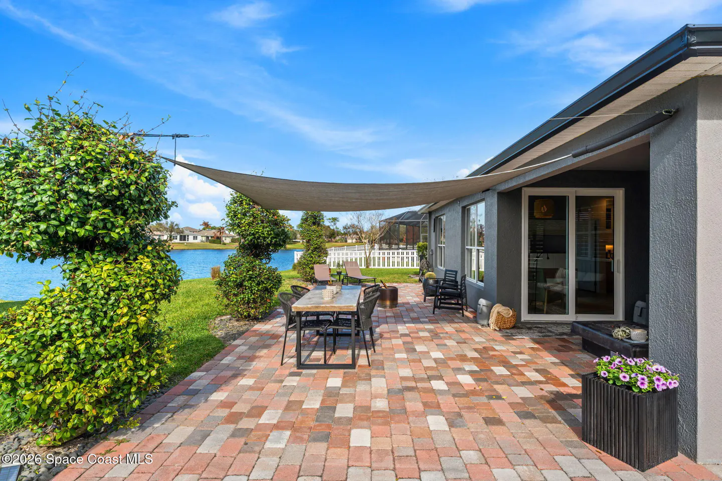 Brick patio with dining table, chairs, and sunshade. Lake view with blue sky. Gray house exterior with sliding glass doors.