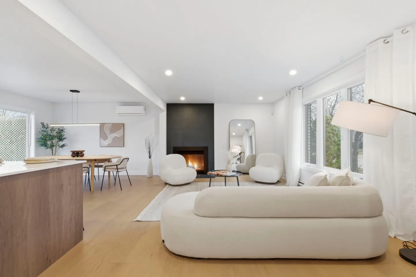 Bright, open-concept living room with white walls, light wood floors, and a black fireplace. White sofa and chairs create a modern, inviting space.