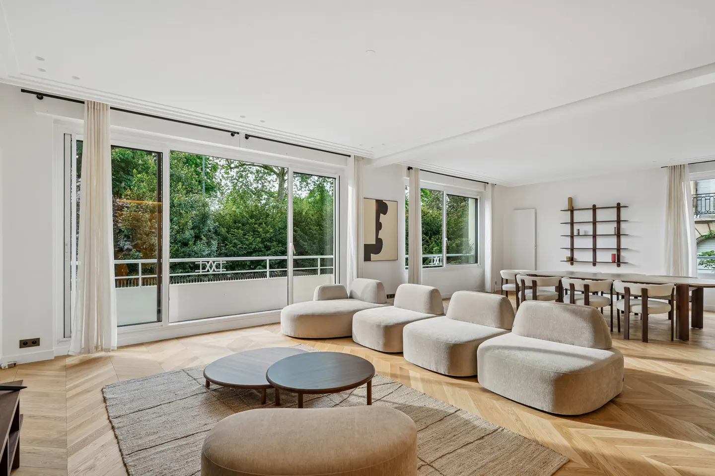 Bright, modern living room with modular beige seating, round tables, and a dining area. Large windows overlook lush greenery.