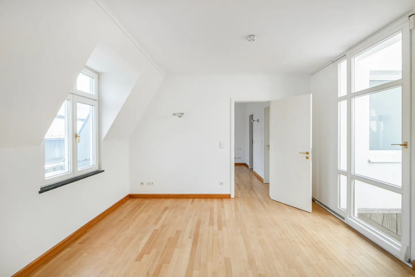 Bright, empty room with wood floors, white walls, and a window. A door is open to another room.