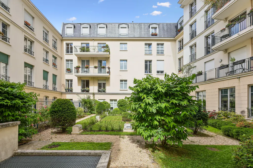 A courtyard garden with gravel paths, green shrubs, and trees surrounded by a multi-story cream apartment building.