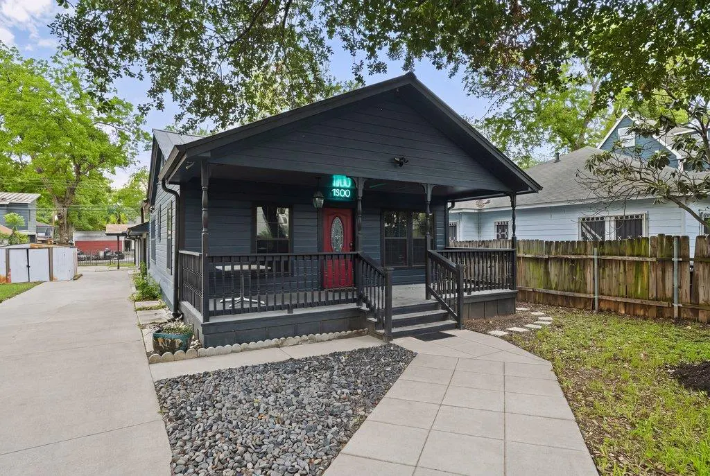 A dark gray house with a red door and a porch with black railings. A sidewalk leads to the house.