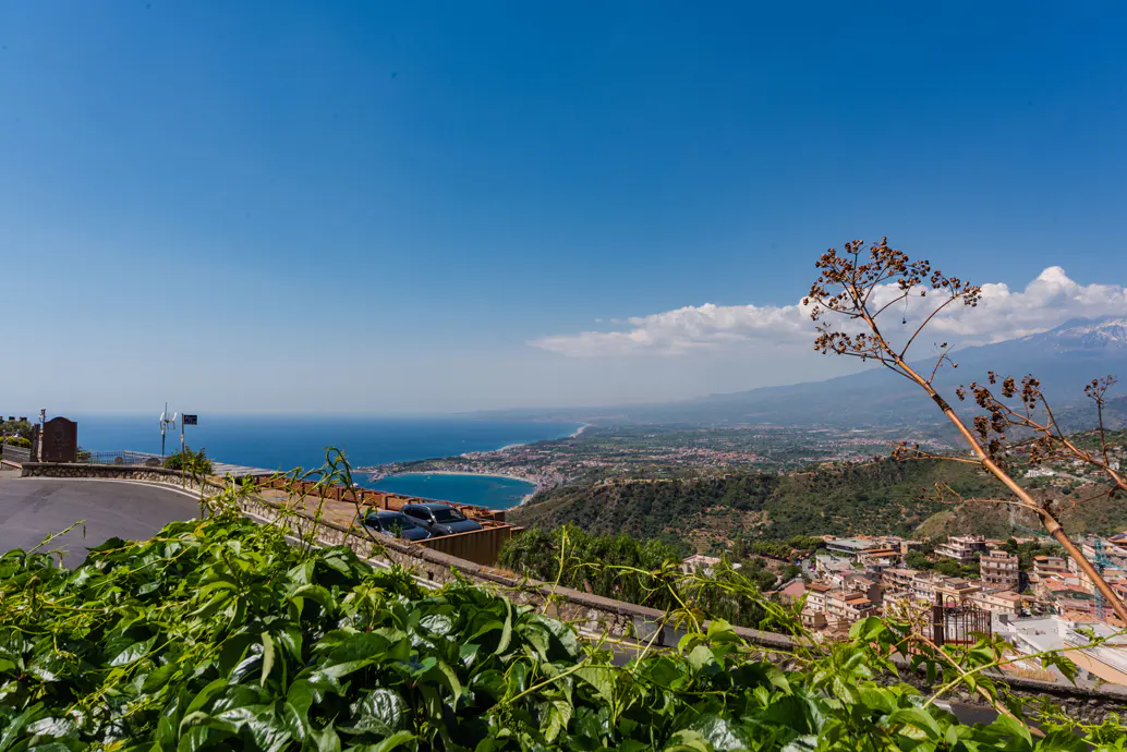 Scenic view from a hillside overlooking a coastal town, blue sea, and distant mountains under a clear sky.