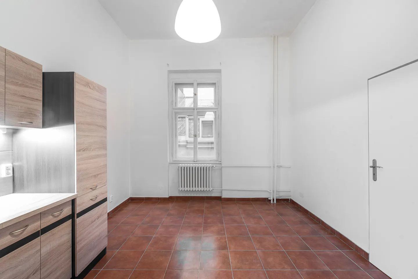 A bright, empty room with brown tile floors, white walls, a window, radiator, and kitchen cabinets.