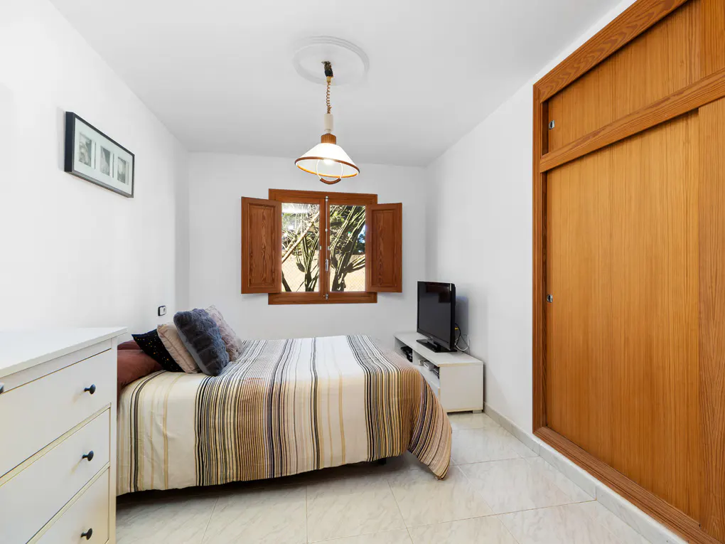 Bedroom with white walls, tiled floor, striped bedspread, and wooden closet. A TV sits on a white stand near a window with wooden shutters.
