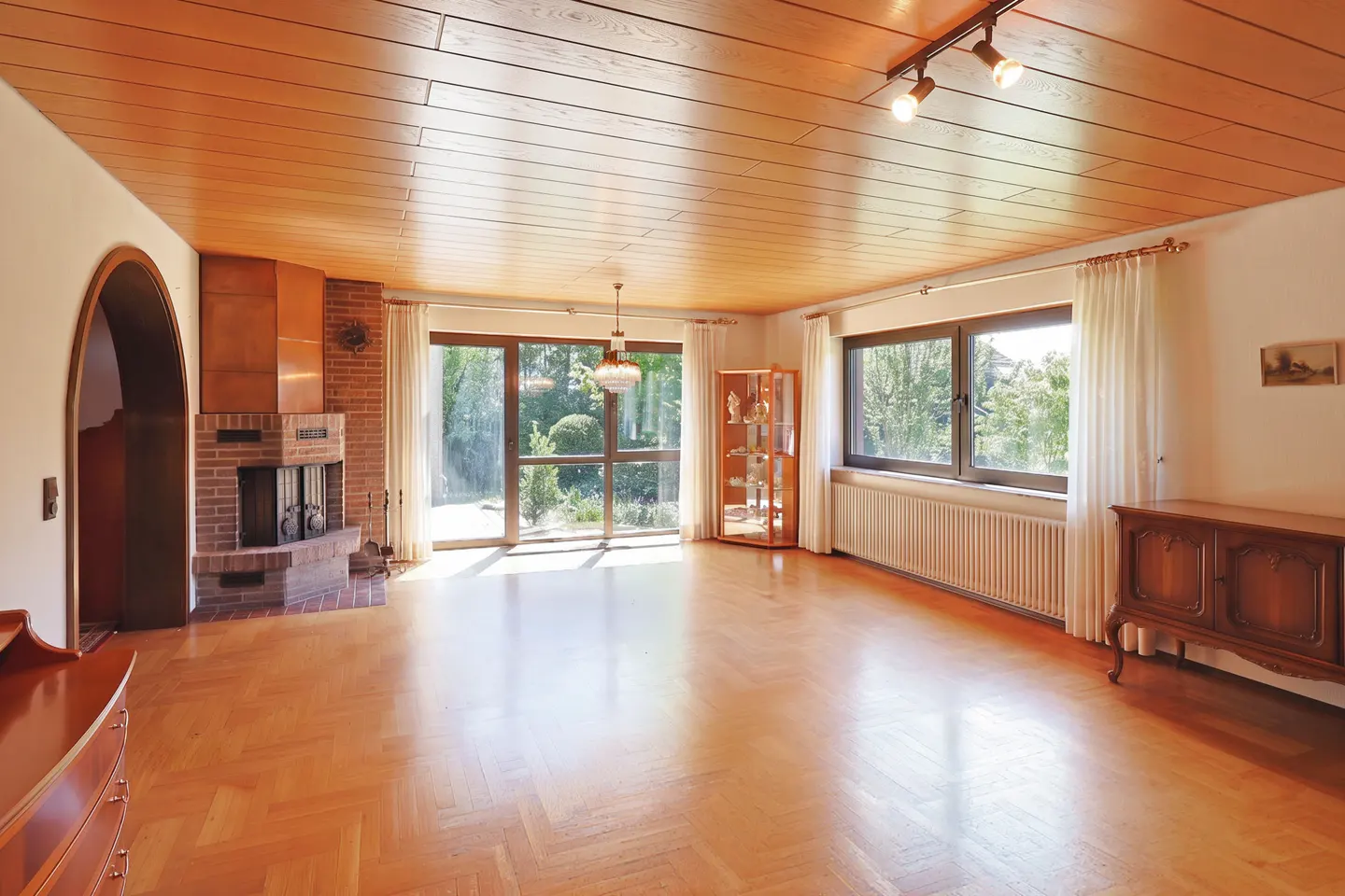 Bright living room with wood floors, a brick fireplace, and large windows overlooking a green garden.