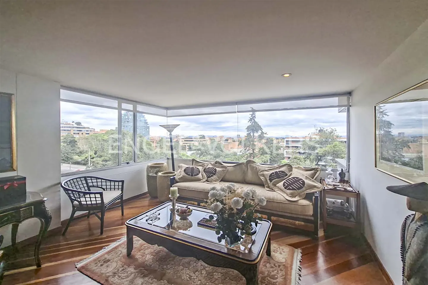 Living room with hardwood floors, a beige sofa with patterned pillows, and a glass coffee table. Large windows overlook a cityscape.
