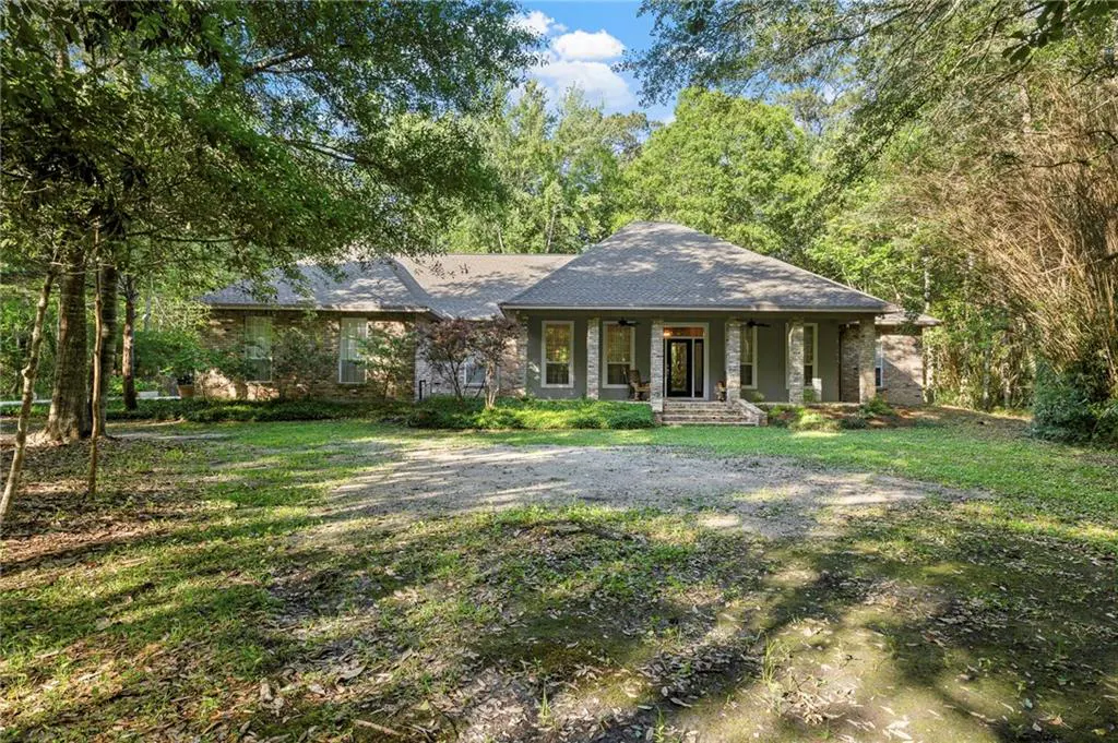 A single-story house with a gray roof, gray siding, and stone pillars, surrounded by trees and a large lawn.