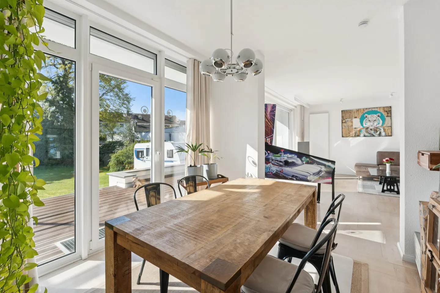 Bright dining room with a wooden table, black chairs, and a modern chandelier. Large windows overlook a green lawn and a white trailer. Artwork and a pinball machine are visible in the background.