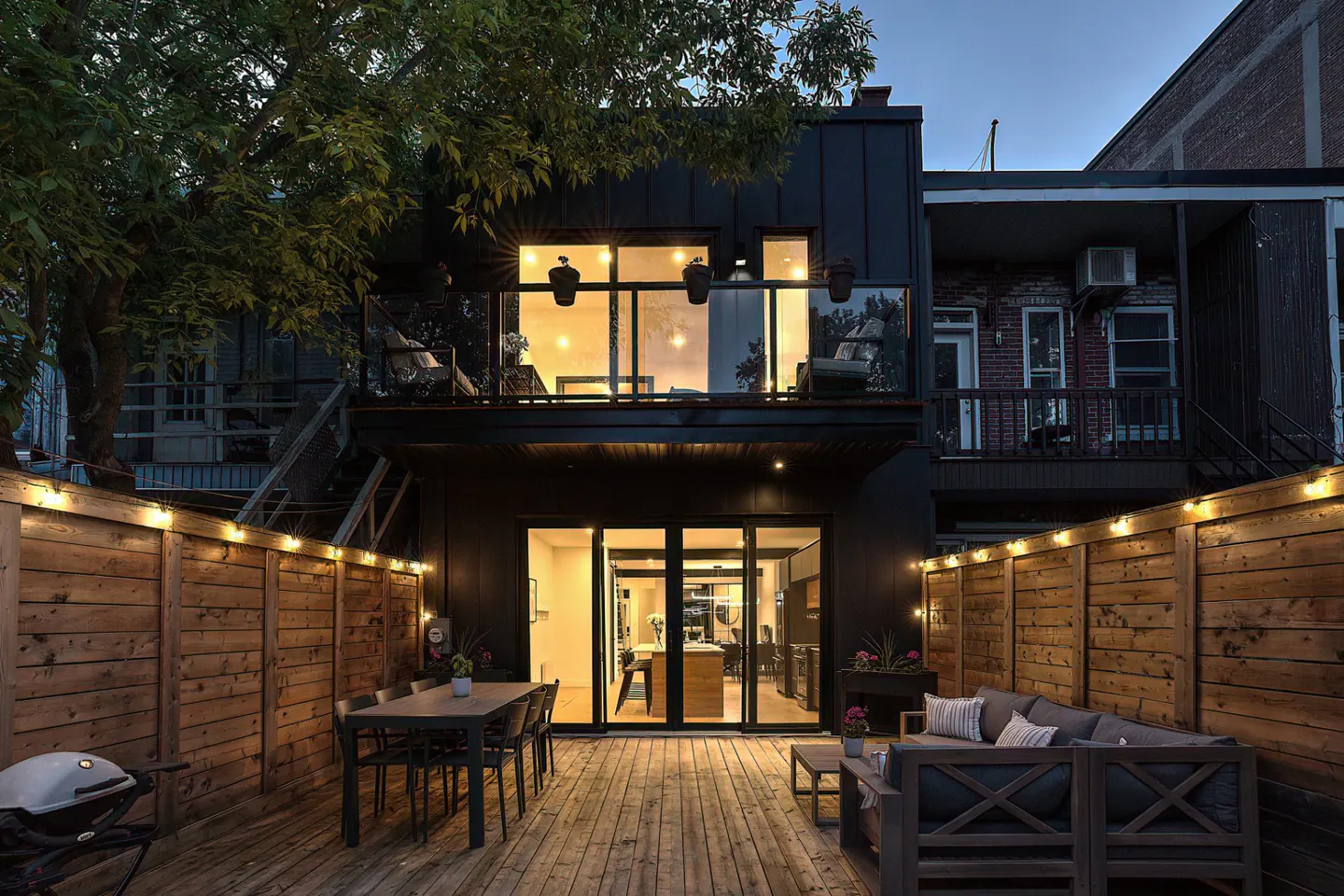 A modern two-story house with a wooden deck, dining table, and outdoor sofa. String lights line the wooden fence.