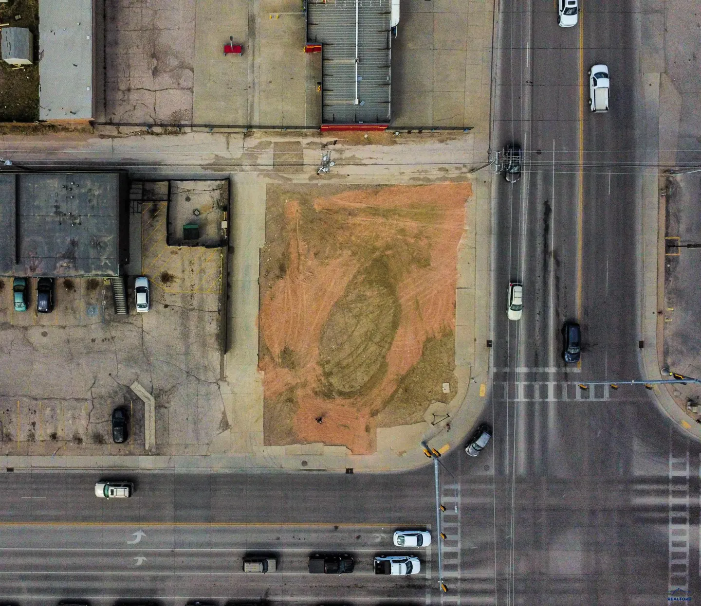 Aerial view of a vacant, dirt-covered urban lot situated between paved roads and commercial buildings with parked cars.