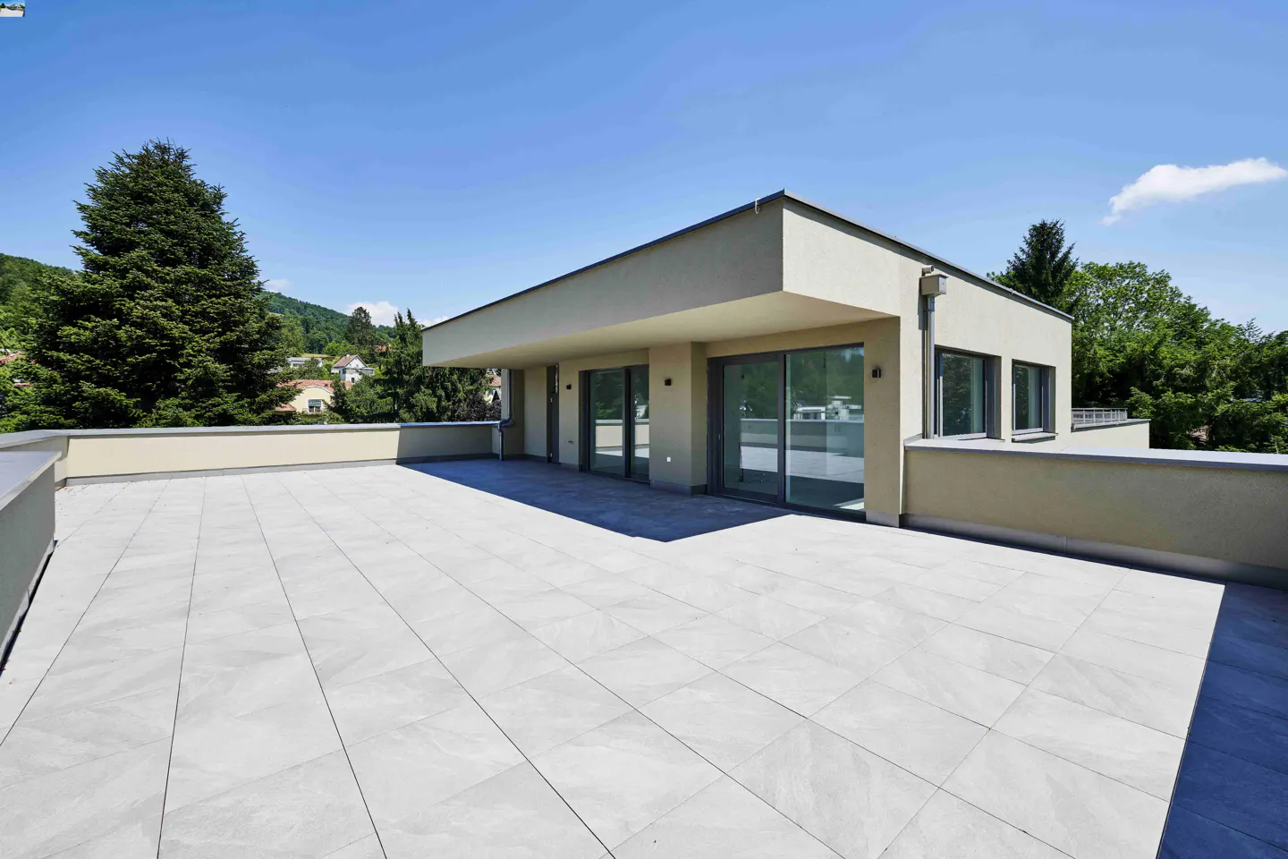 Spacious rooftop terrace with light grey tiles, modern beige building exterior, large glass doors, and a clear blue sky.