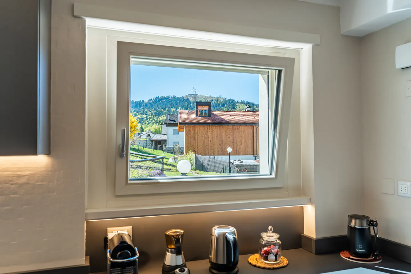 A kitchen window offers a view of a house and a mountain under a blue sky. Coffee makers sit on the counter below.