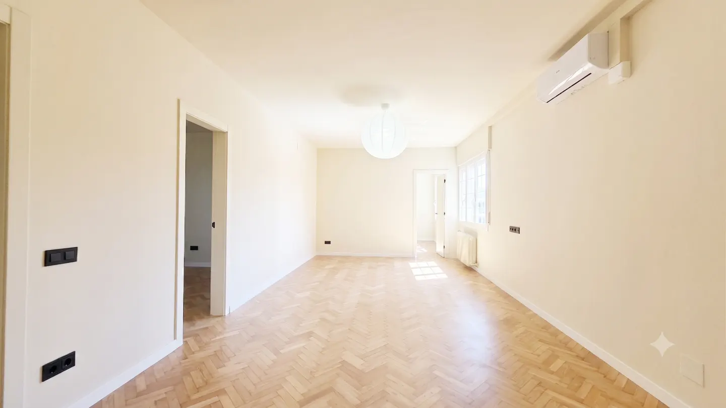 Bright, empty room with cream walls, herringbone wood floors, a white globe pendant light, and a wall-mounted AC unit.