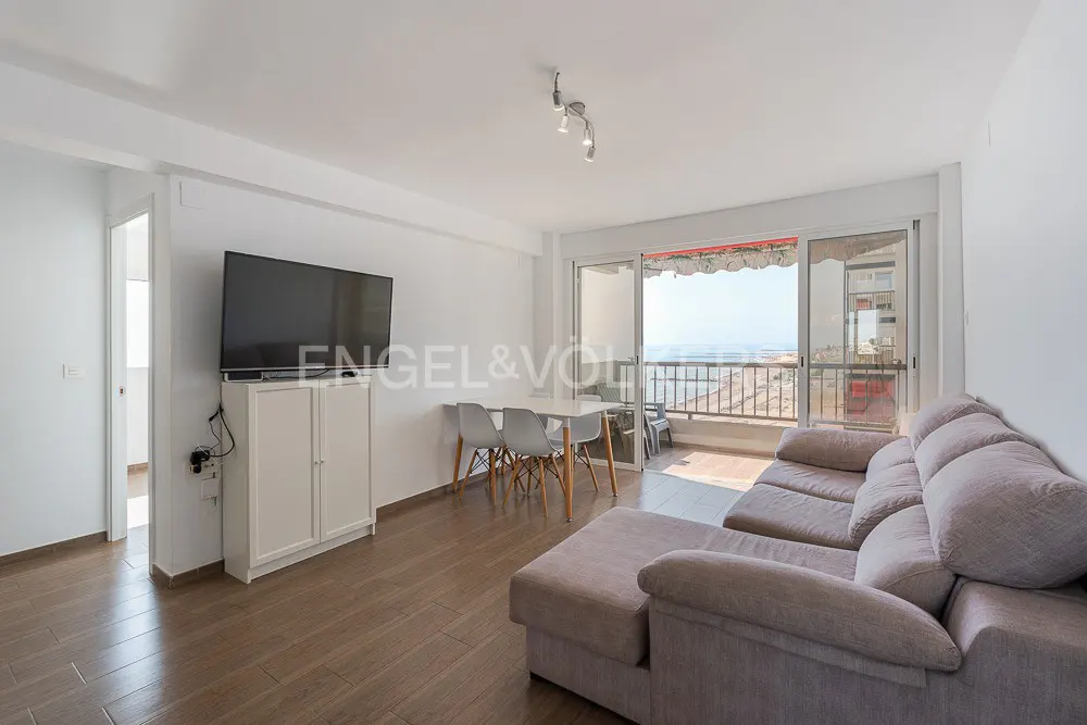 Bright living room with a gray sofa, white table and chairs, TV, and balcony overlooking the ocean.