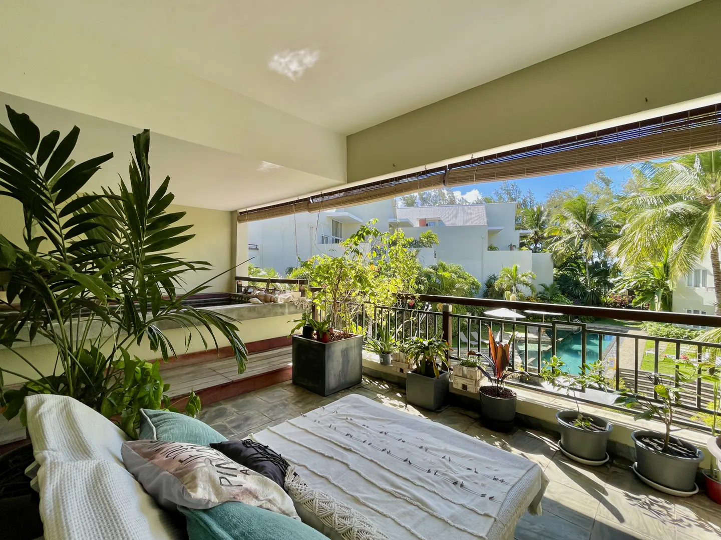 Balcony view with plants, a daybed, and a jacuzzi. Outside, there's a pool, palm trees, and white buildings.