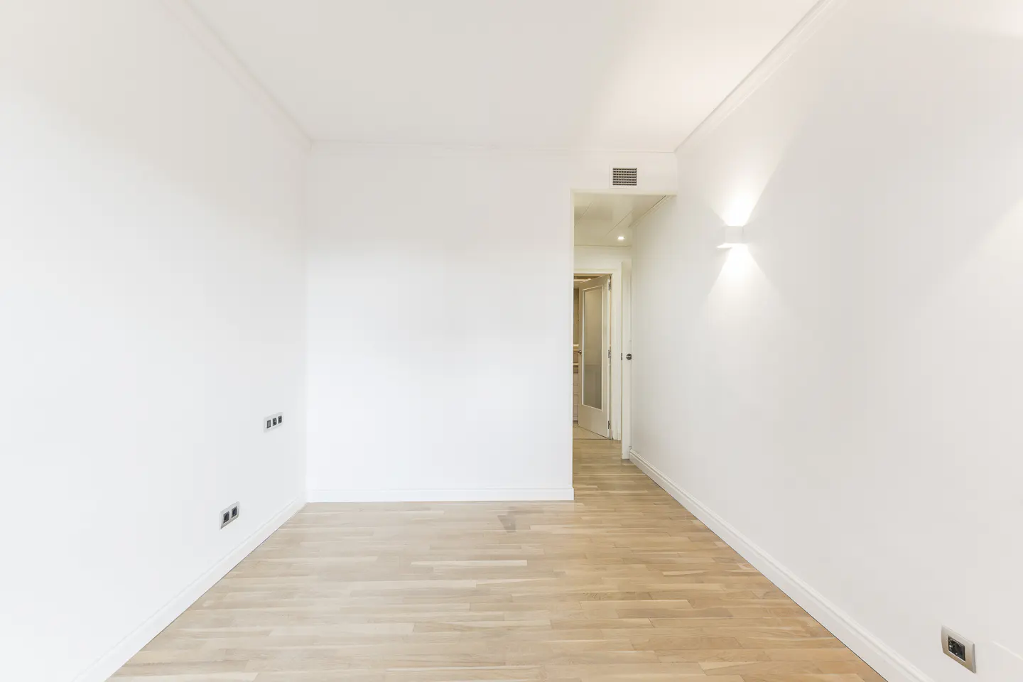Empty room with light wood floors and white walls. A doorway leads to another room. Wall sconce provides light.