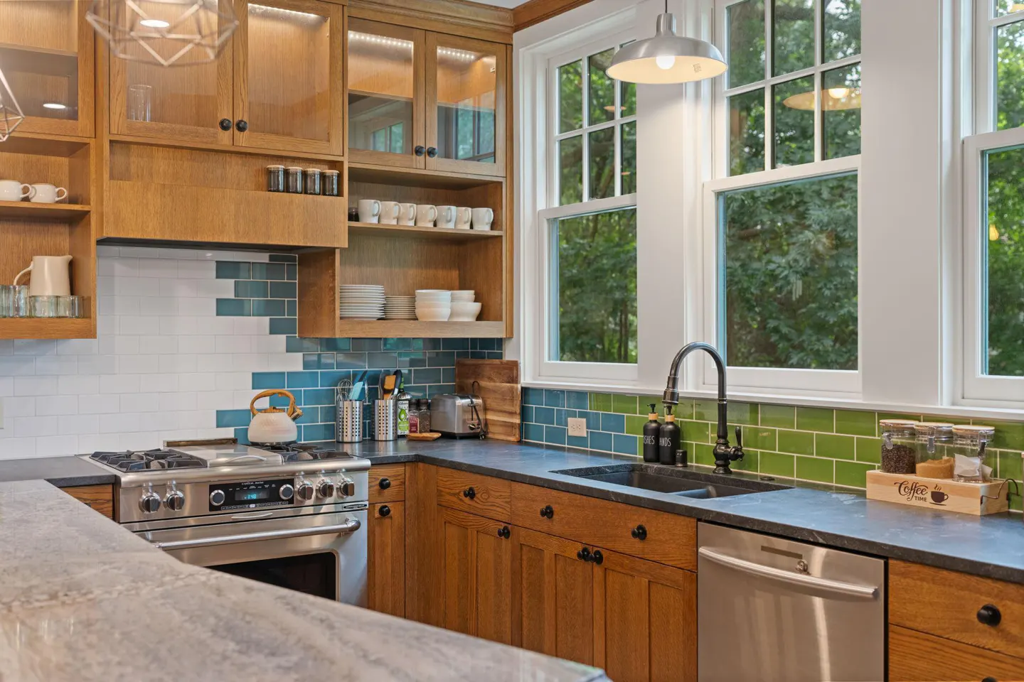 A well-lit kitchen with oak cabinets, stainless steel appliances, and a blue and green tiled backsplash. Large windows overlook a green outdoor space.