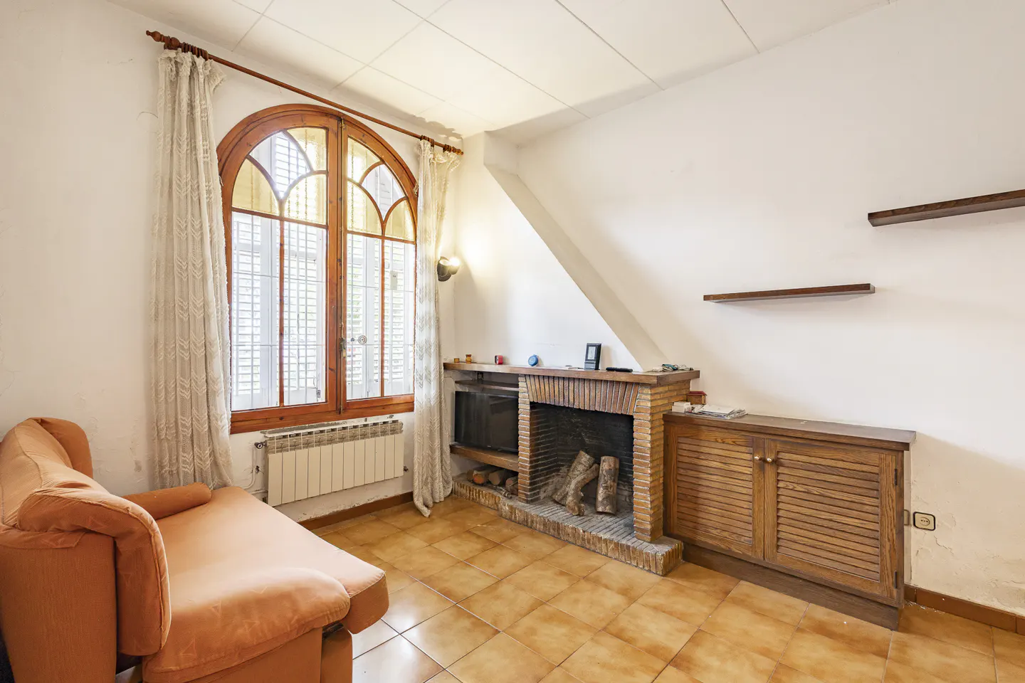 Living room with orange chaise lounge, arched window with curtains, brick fireplace, and brown tiled floor.