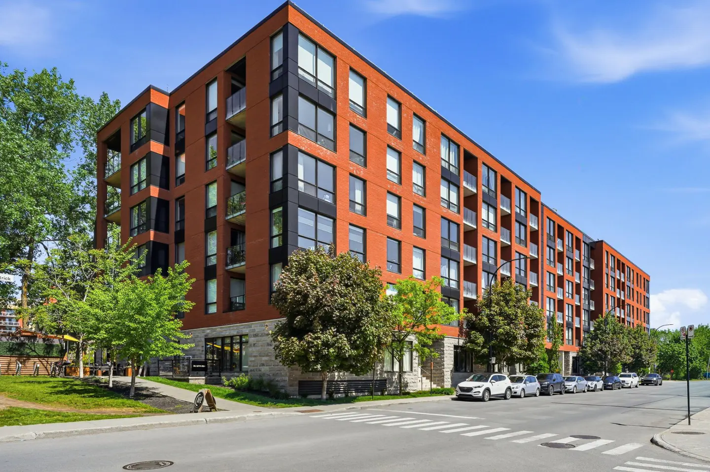 Modern multi-story apartment building with red brick and black accents, featuring balconies and street-level parking.