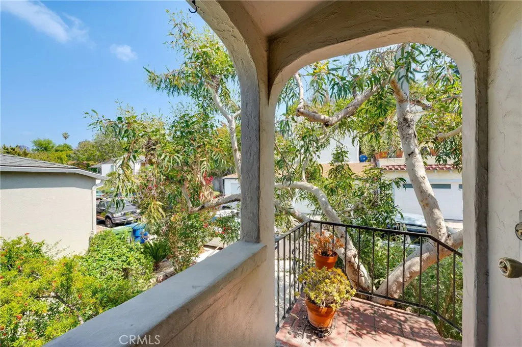 View from an arched balcony with potted plants, overlooking a residential street with trees and houses on a sunny day.